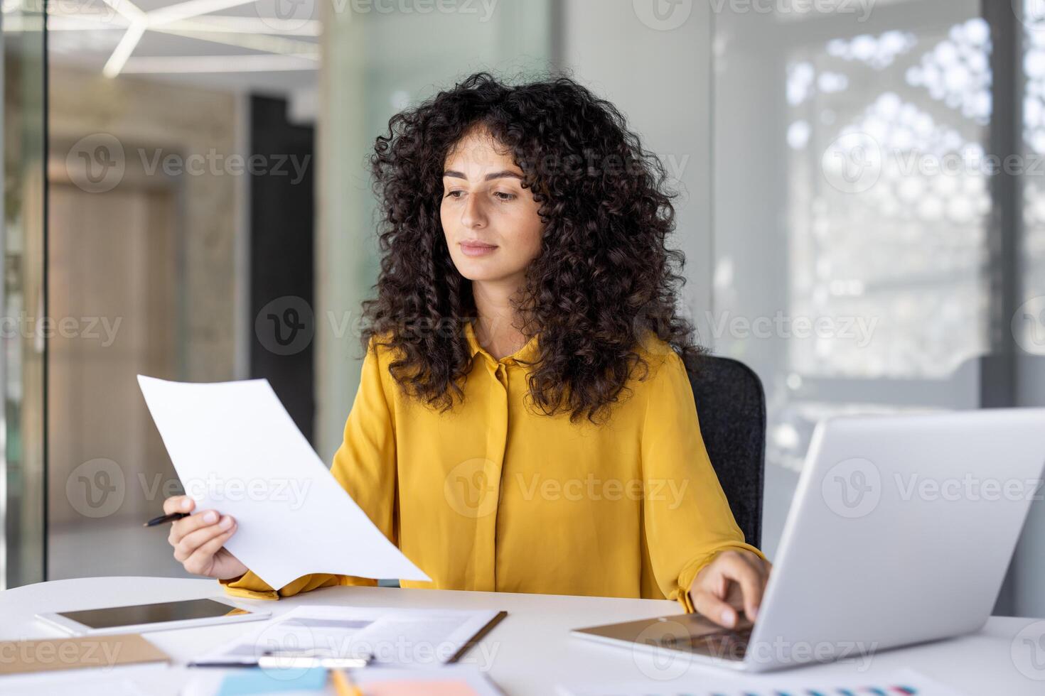 Serious thinking woman holds a document in her hands, reads and checks statistical indicators. Businesswoman at work inside the office uses a laptop, among bills, reports and documents. photo