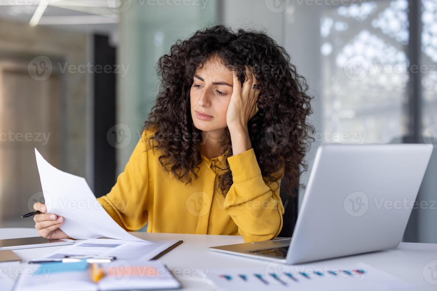Hispanic businesswoman sits at desk with laptop, reviewing documents with concentrated expression. Image conveys stress, analysis, and multitasking in workplace setting. photo