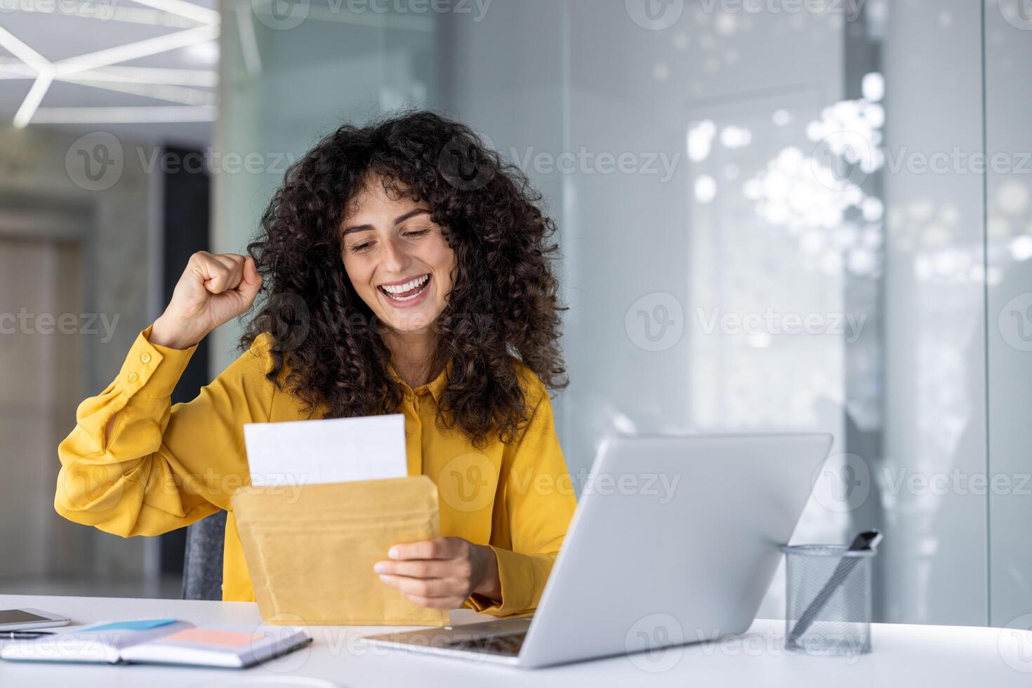 Happy woman in yellow shirt rejoicing positive news at desk with laptop. She holds envelope with excited expression, symbolizing career achievement and satisfaction at work. Modern workspace ambiance photo
