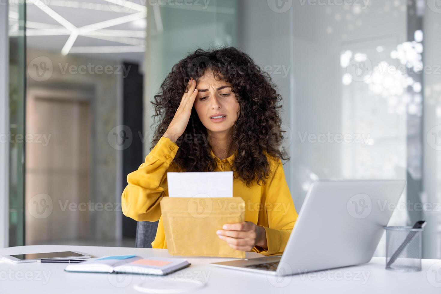 Stressed woman receiving bad news from envelope in office setting. Expression of worry while seated at desk with laptop, phone, and various office essentials. Modern professional environment. photo