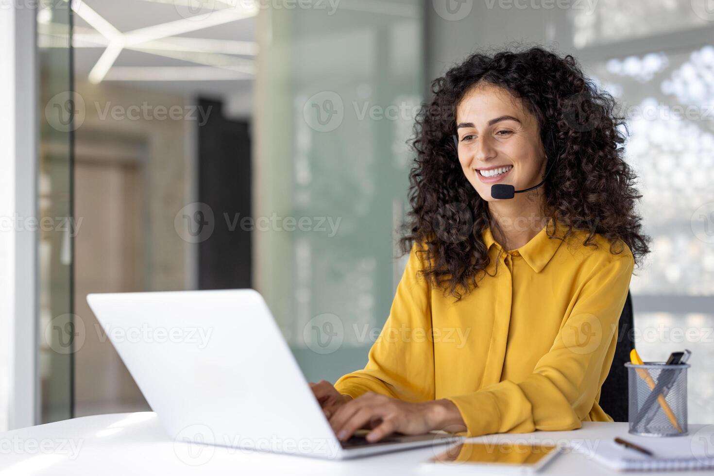 Joyful customer support and consulting service worker with headset smiling, using laptop for call. Woman inside office, remotely helping customers, supporting. photo