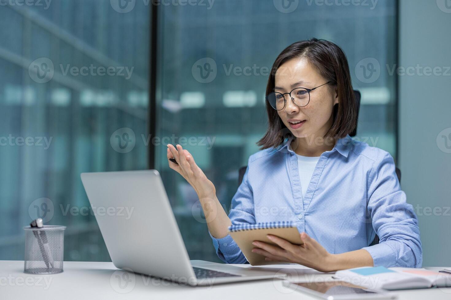 Online business meeting, young woman talking to colleagues remotely and writing data in notebook, using laptop for call, Asian businesswoman smiling happily watching online training course. photo
