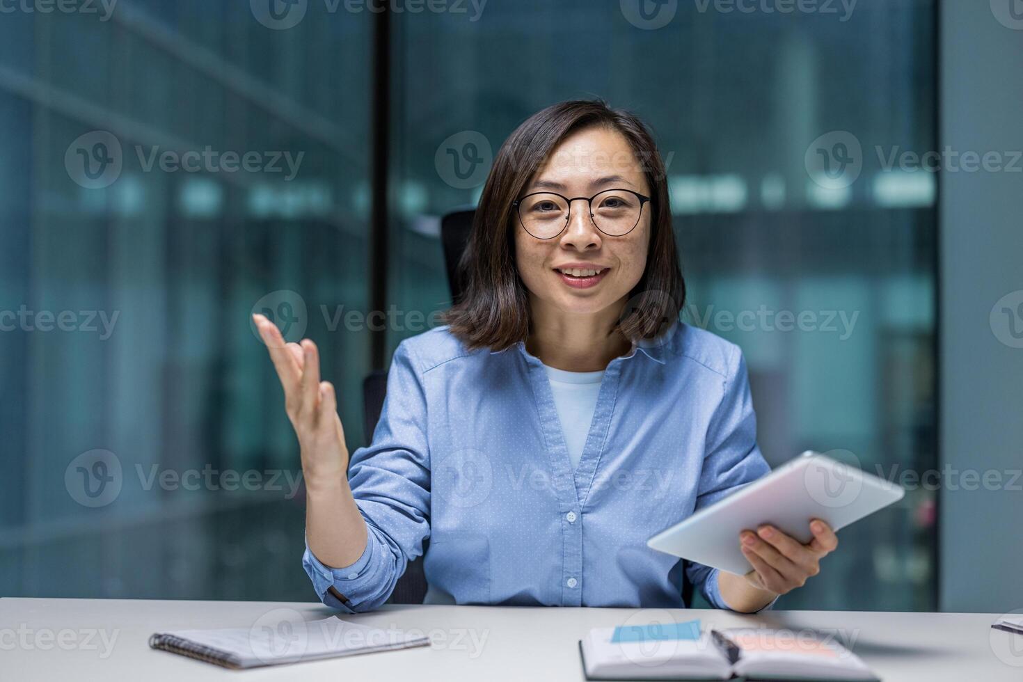 Joyful Asian woman with tablet computer in hands sitting at desk inside office. Woman smiling and looking at camera, webcam view, online presentation business meeting. photo