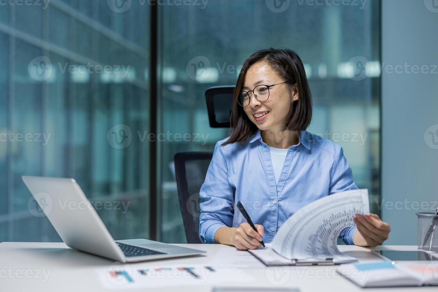Joyful and successful Asian woman doing paperwork inside the office, businesswoman financier preparing reports and contracts, filling out economic indicators and tax forms, using laptop. photo