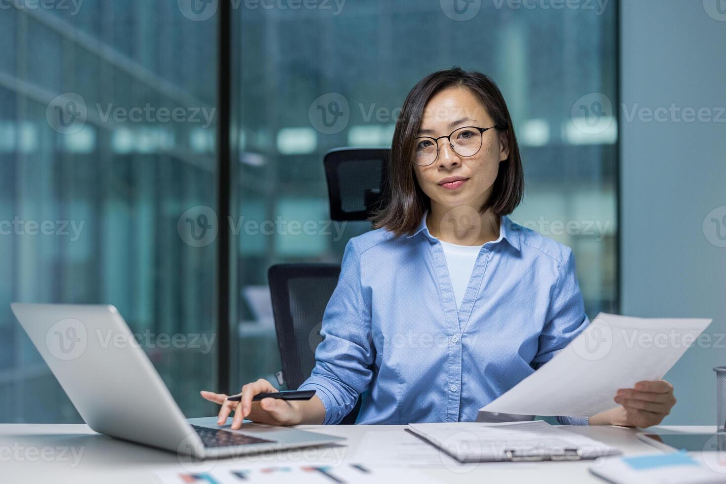 Asian businesswoman working with documents and a laptop in a modern office setting. She holds a professional demeanor, clearly showcasing her focus and dedication to task. photo