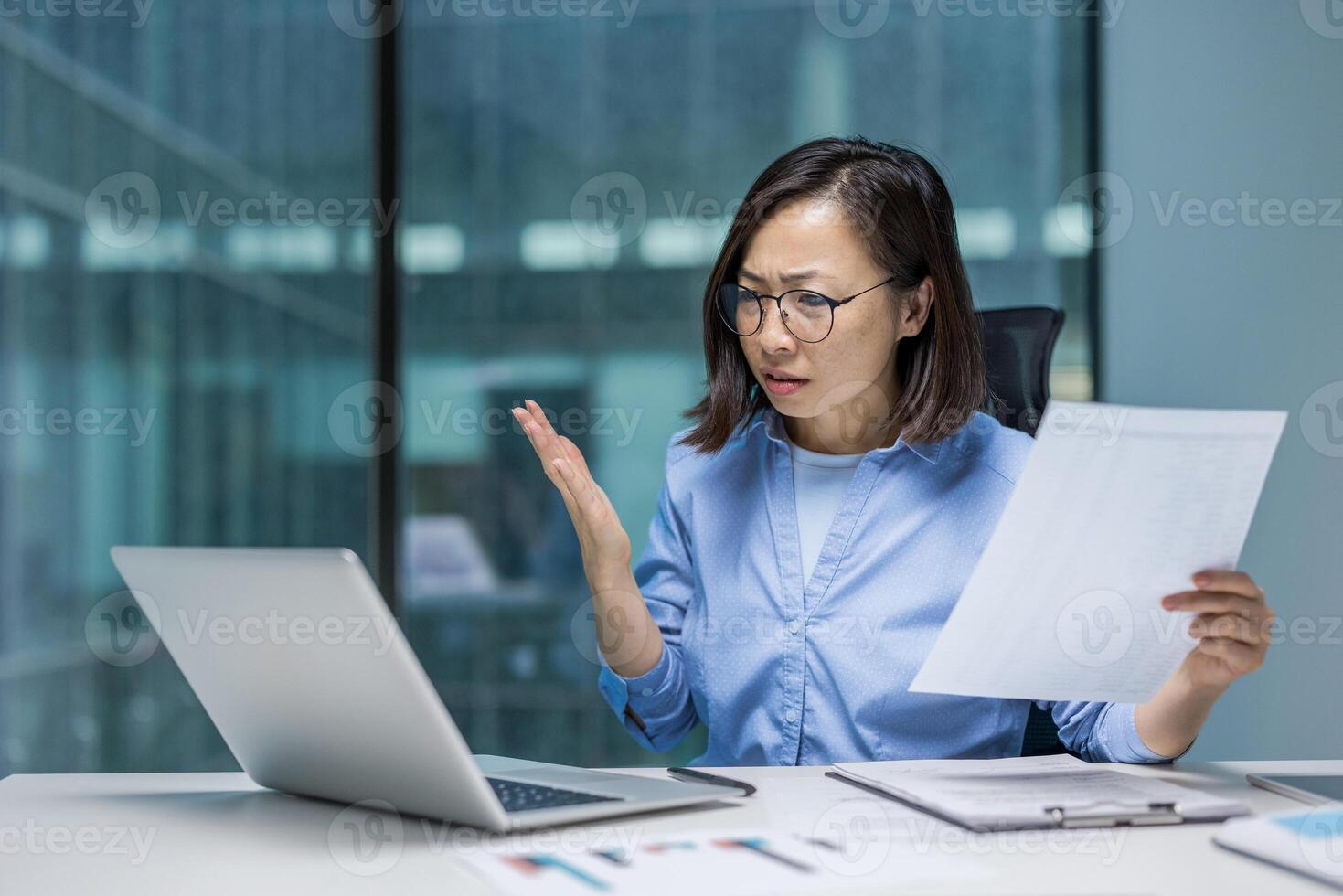 Asian businesswoman expressing frustration reviewing financial documents in a modern office. Holding a sheet of paper, she looks at her laptop with a perplexed expression. photo