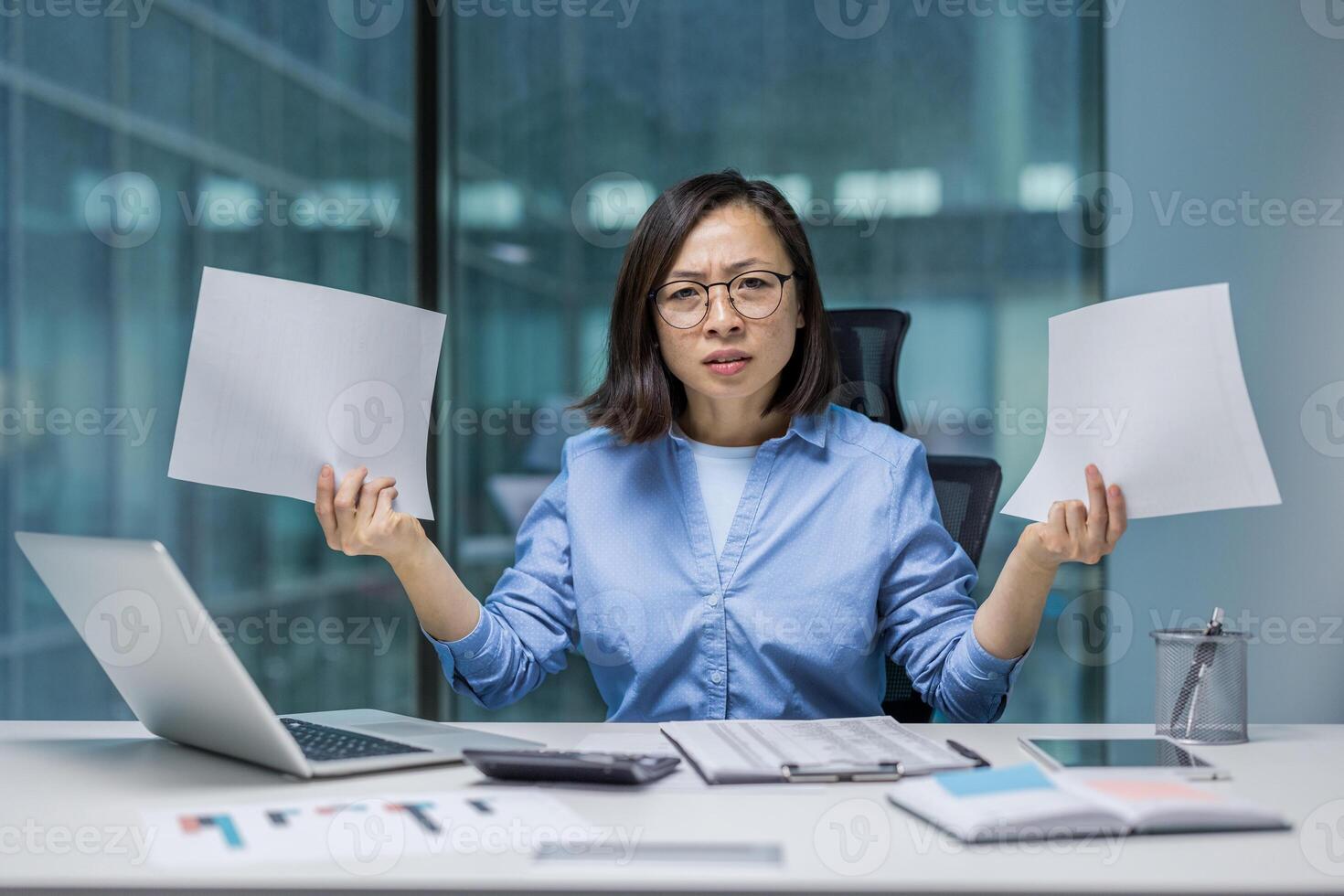 Asian businesswoman expressing frustration holding documents at her desk in a modern office. The scene depicts challenges faced in workplace, decision-making, and paperwork management in office photo