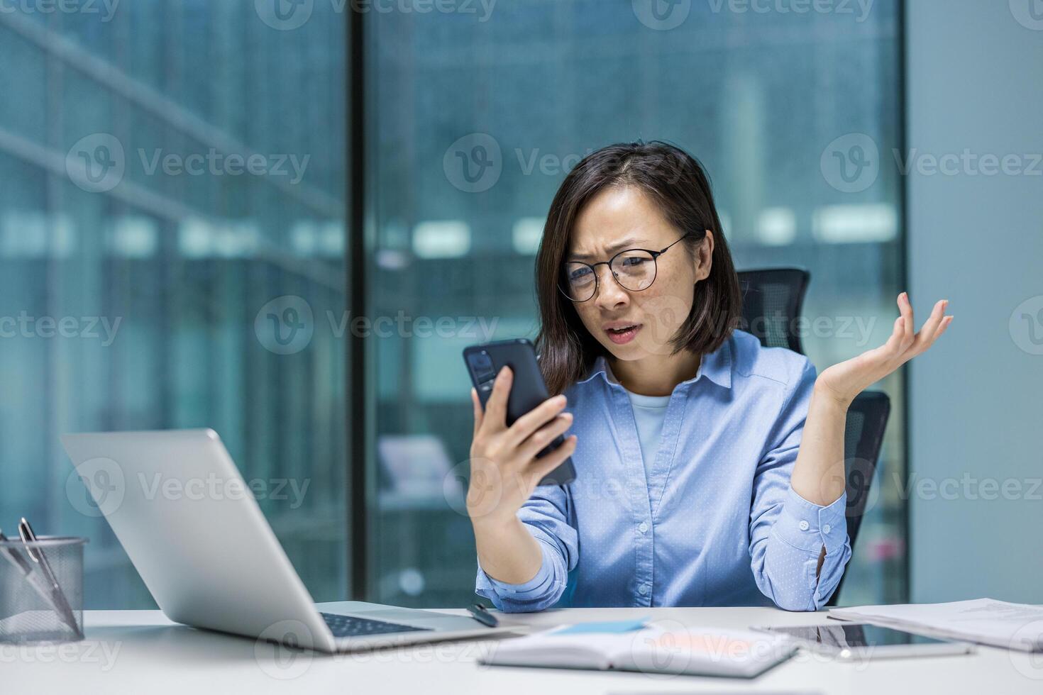Asian businesswoman experiencing frustration as she examines her smartphone. Seated at a desk with a laptop and documents, her expression reflects concern or confusion. photo