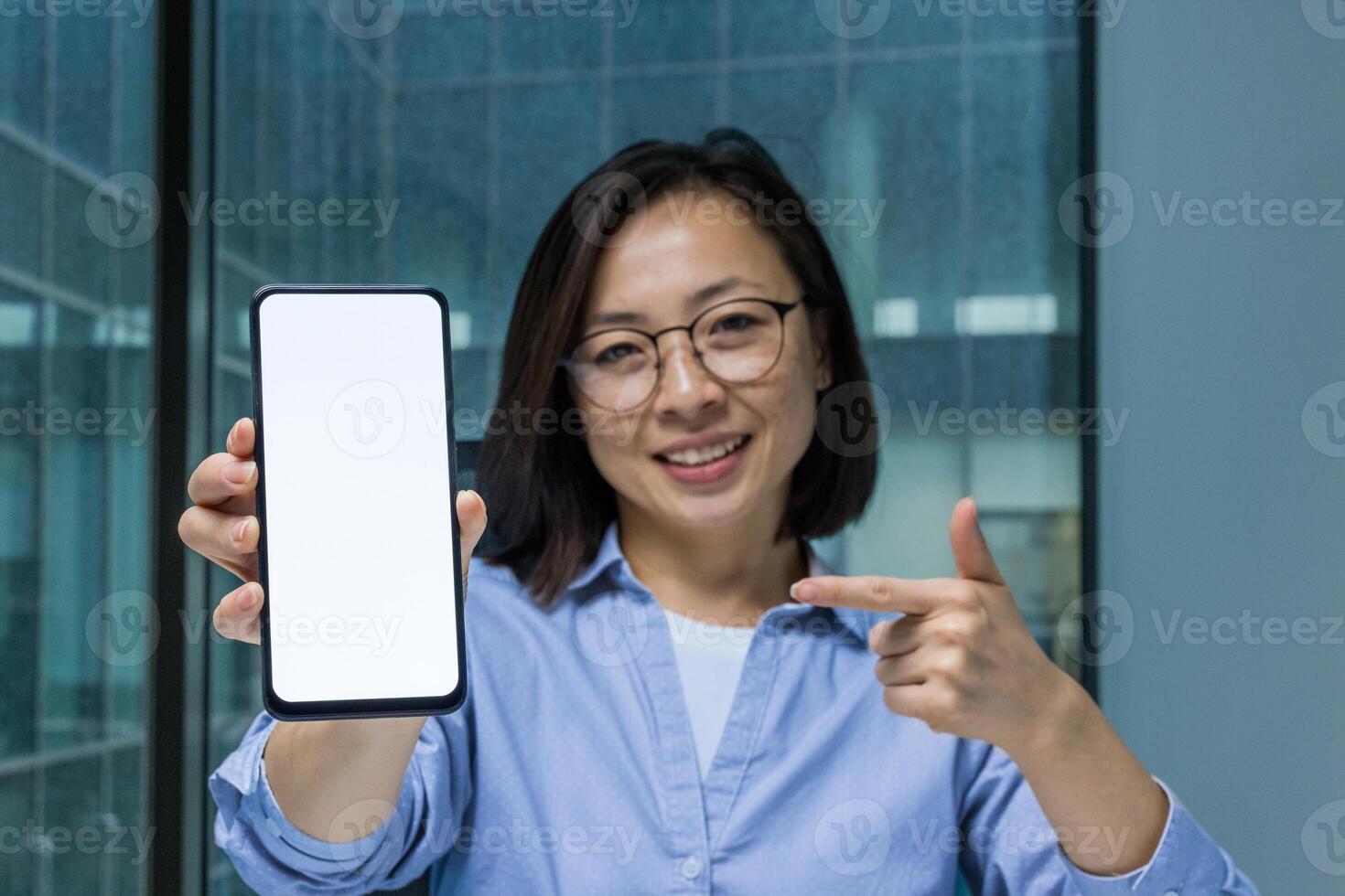 Asian businesswoman holding a smartphone with a blank screen, pointing towards it and smiling. Perfect for representing technology, business presentations, or mockups for app design and development. photo