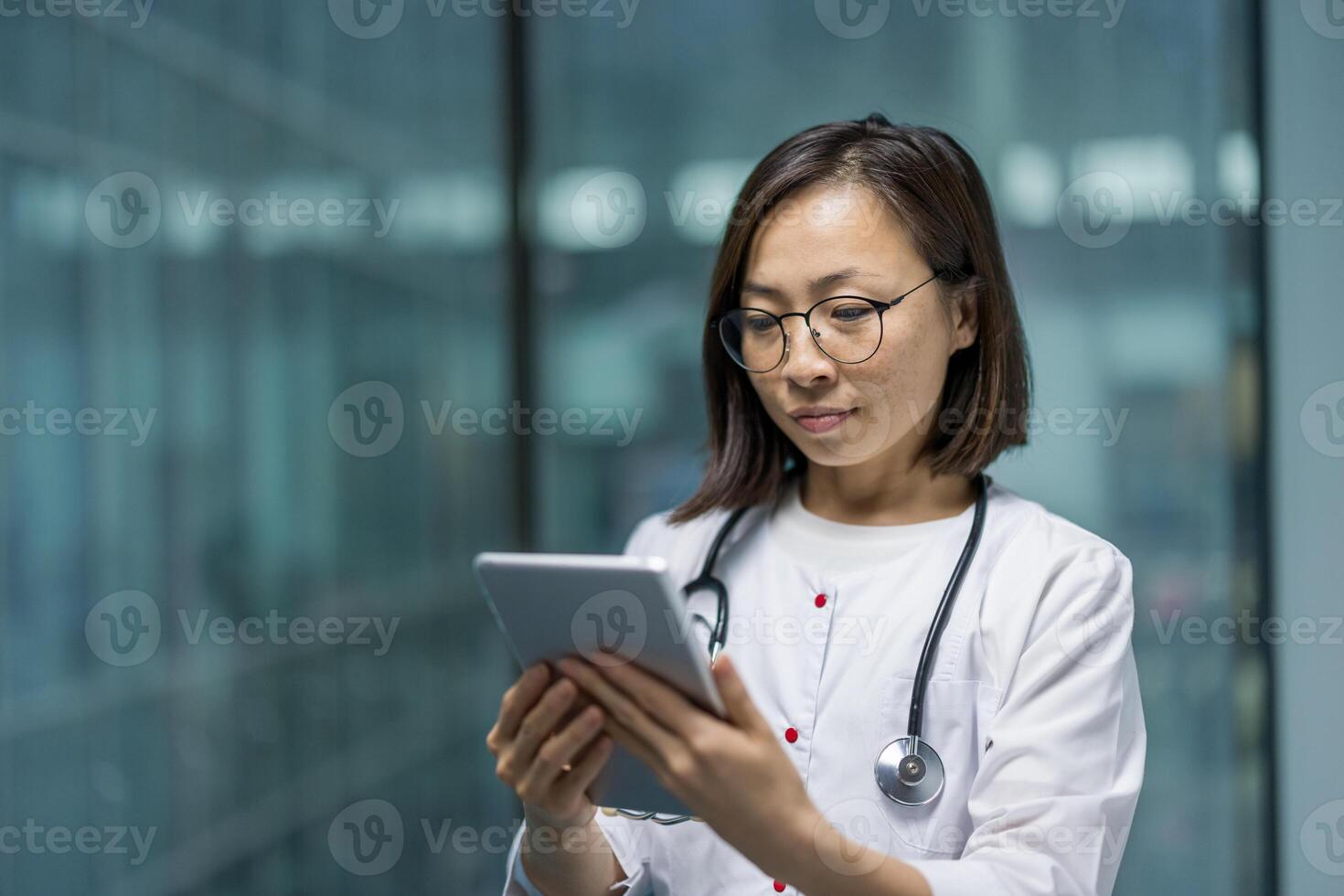 Asian woman doctor wearing eyeglasses and a stethoscope, interacting with a digital tablet to review medical information and notes. photo