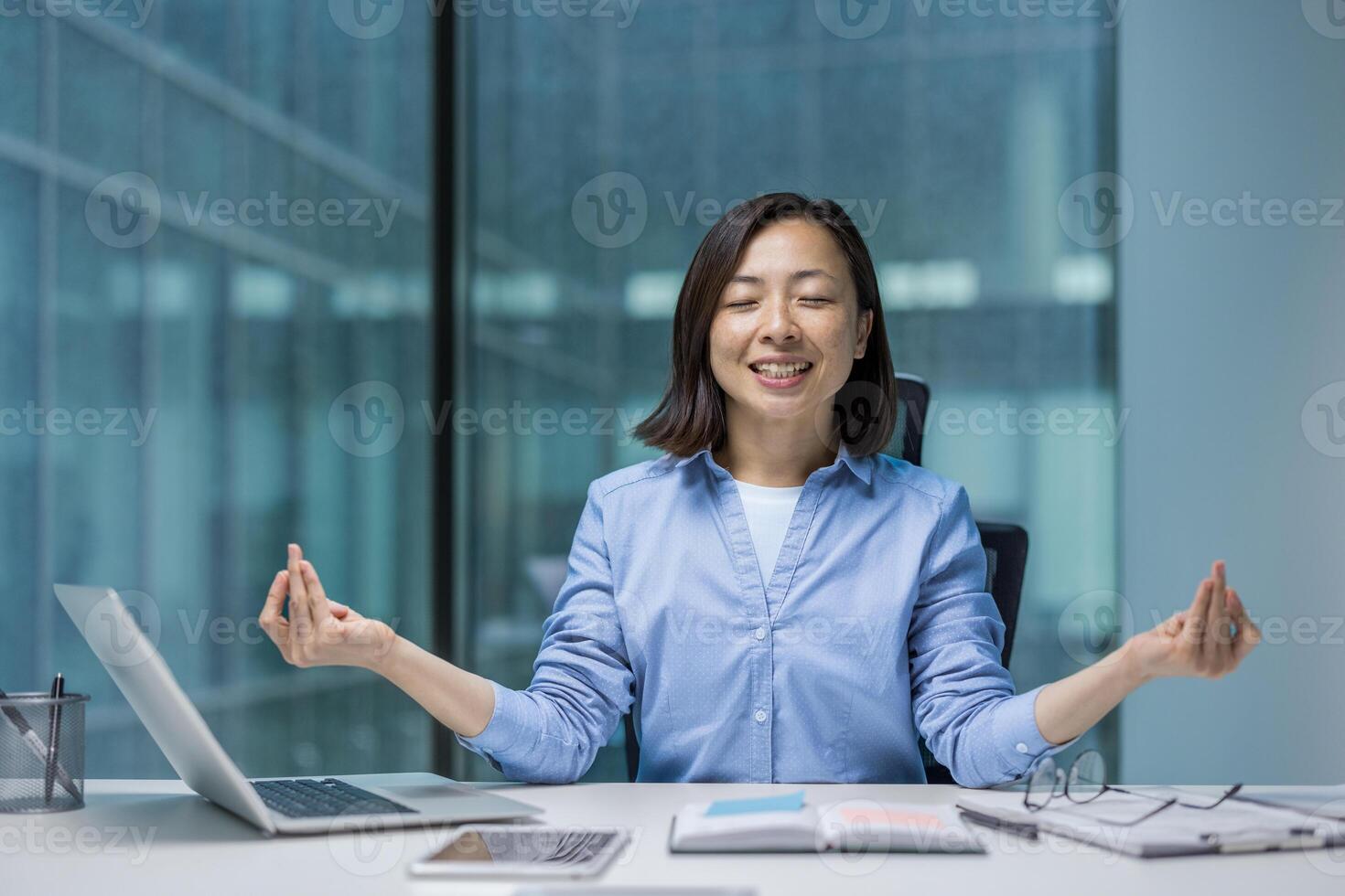 Young joyful Asian woman meditating, resting and relaxing in lotus position. Woman smiling contentedly doing breathing exercises. Businesswoman inside office at workplace, calm and focused. photo
