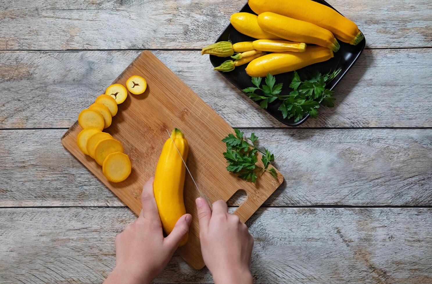 Two hands are slicing a yellow squash into round pieces on a wooden cutting board. The scene includes other vegetables like lettuce, garlic, cauliflower, and a carrot, set on a dark countertop photo