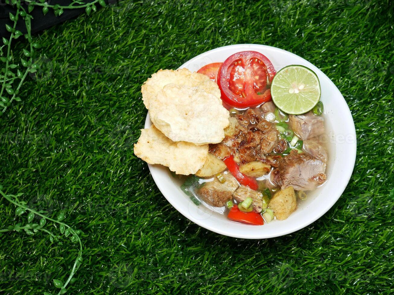 Beef Soup with sliced Beef, Potatoes, Tomatoes, Lime, crackers, sprinkled with spring onions, and fried onions. Served in a white bowl with a green synthetic grass background. photo