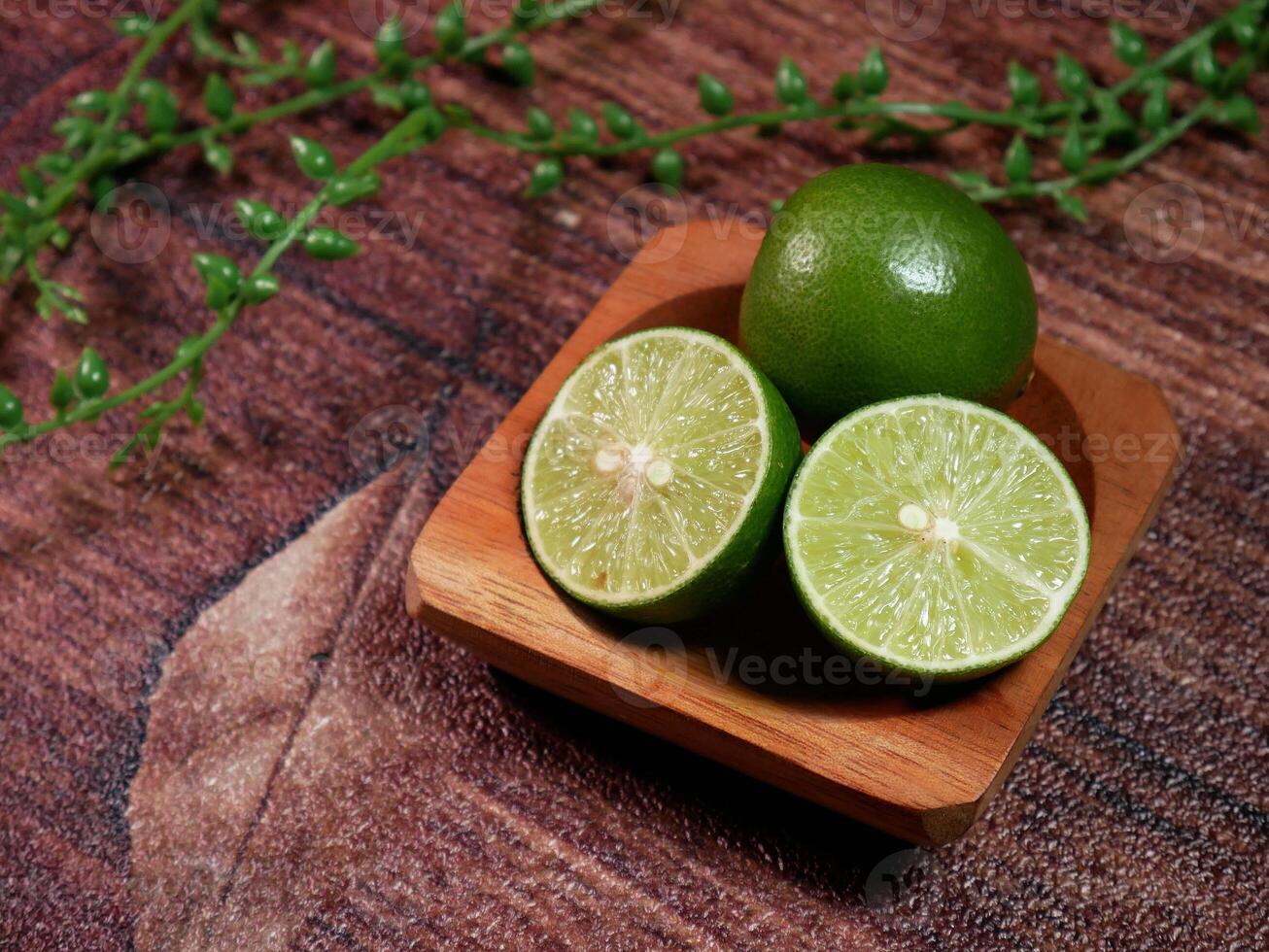 Closeup Fresh limes, consisting of one whole lime and two slices, neatly arranged on a small wooden plate. With brown background and decorative leaves enhance the natural. photo