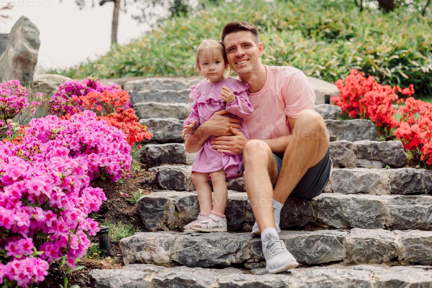 Happy father and daughter enjoying time together, sitting on stone steps surrounded by vibrant flowers in a spring garden. photo