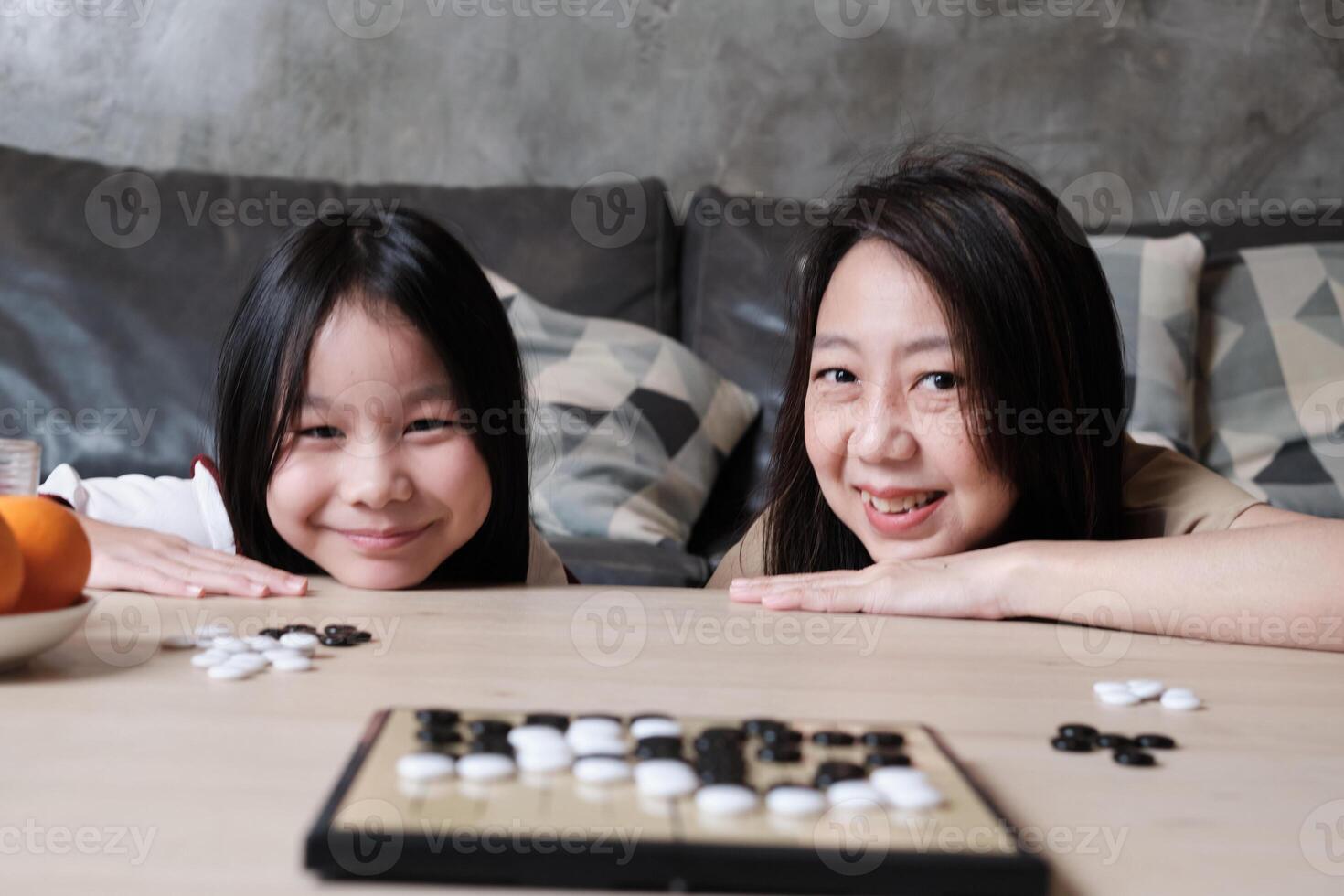 Portrait of Asian family, mum, and kid fun playing Go, checkers board game on table in home living room with, happy weekend times relaxing hobby, smile and look at camera together. photo