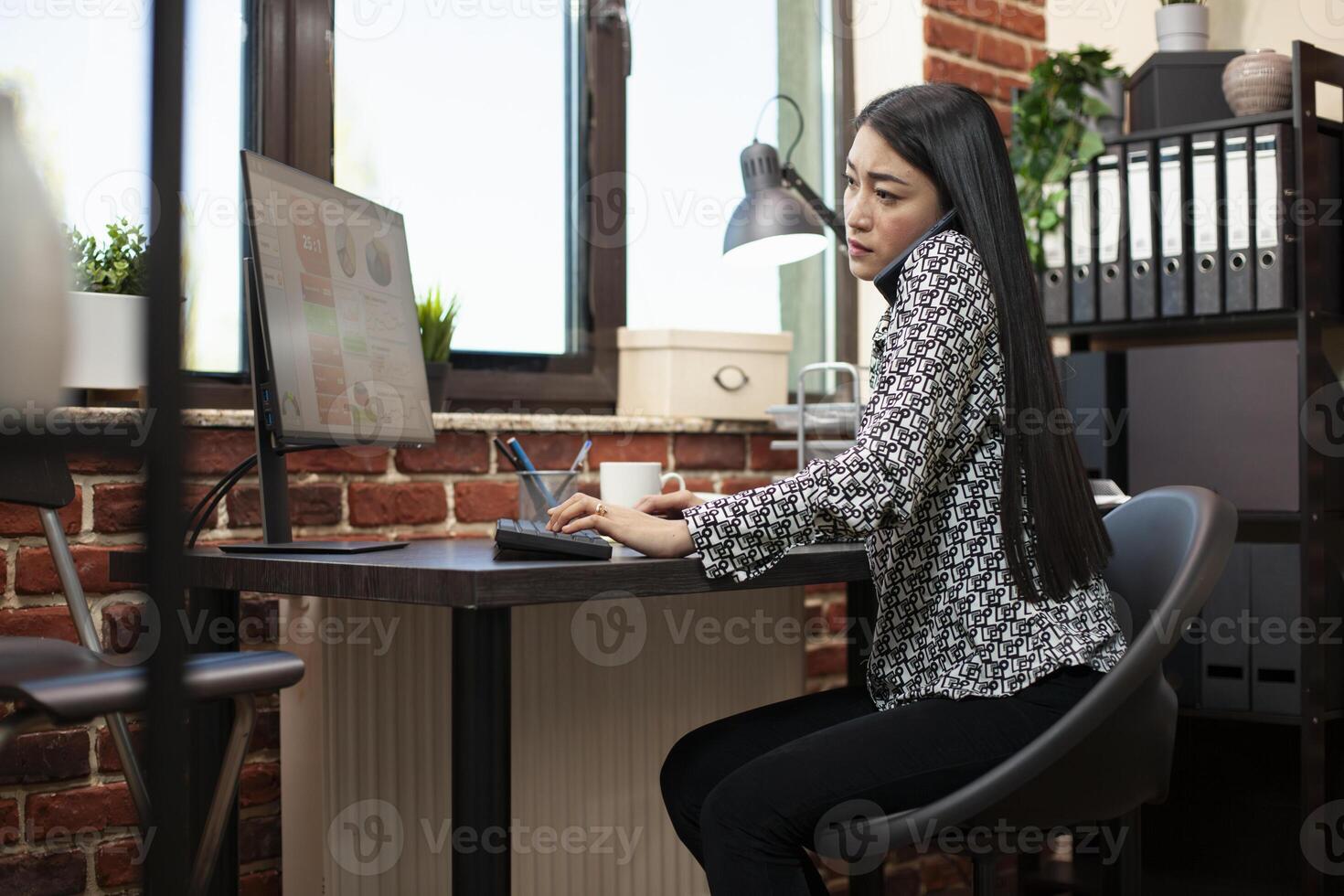 Female manager multitasks during phone call with client, entering details on computer in startup office. Asian woman seated at her desk, using keyboard and speaking with colleague on mobile device. photo