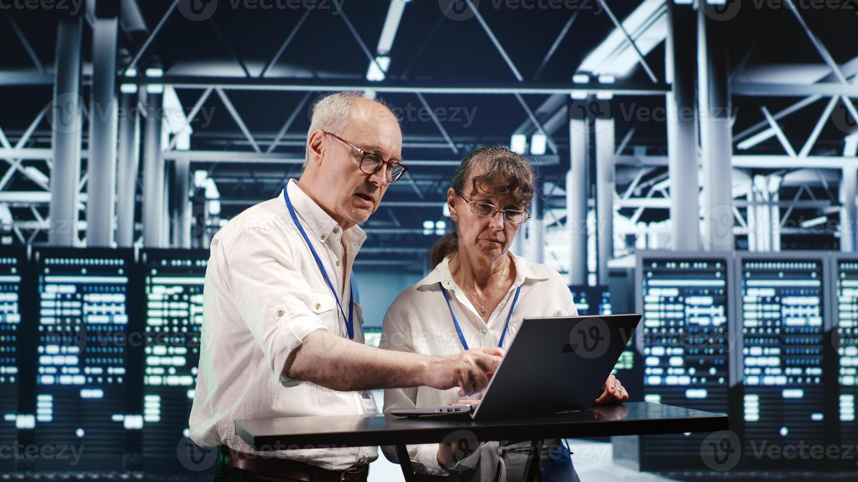 Senescent colleagues looking around high tech data center, using laptop to crosscheck disaster recovery plan and assess server machines in need of replacement, preventing glitches photo