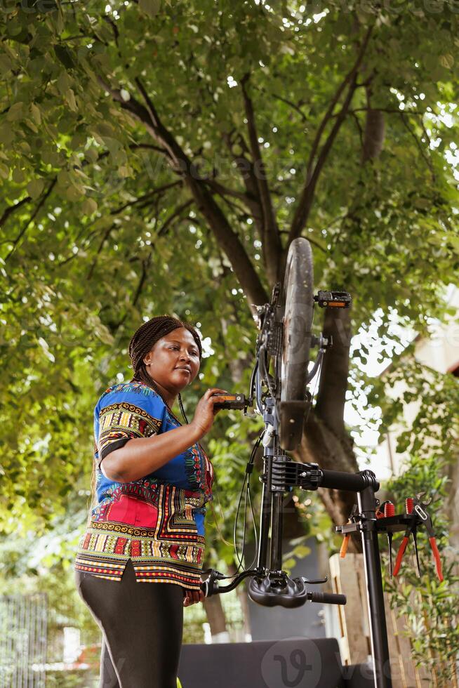 Healthy enthusiastic african american woman mending her own bicycle in the yard doing annual outdoor vehicle maintenance using expert tools. Inspecting and fixing bike pedal as summer hobby. photo