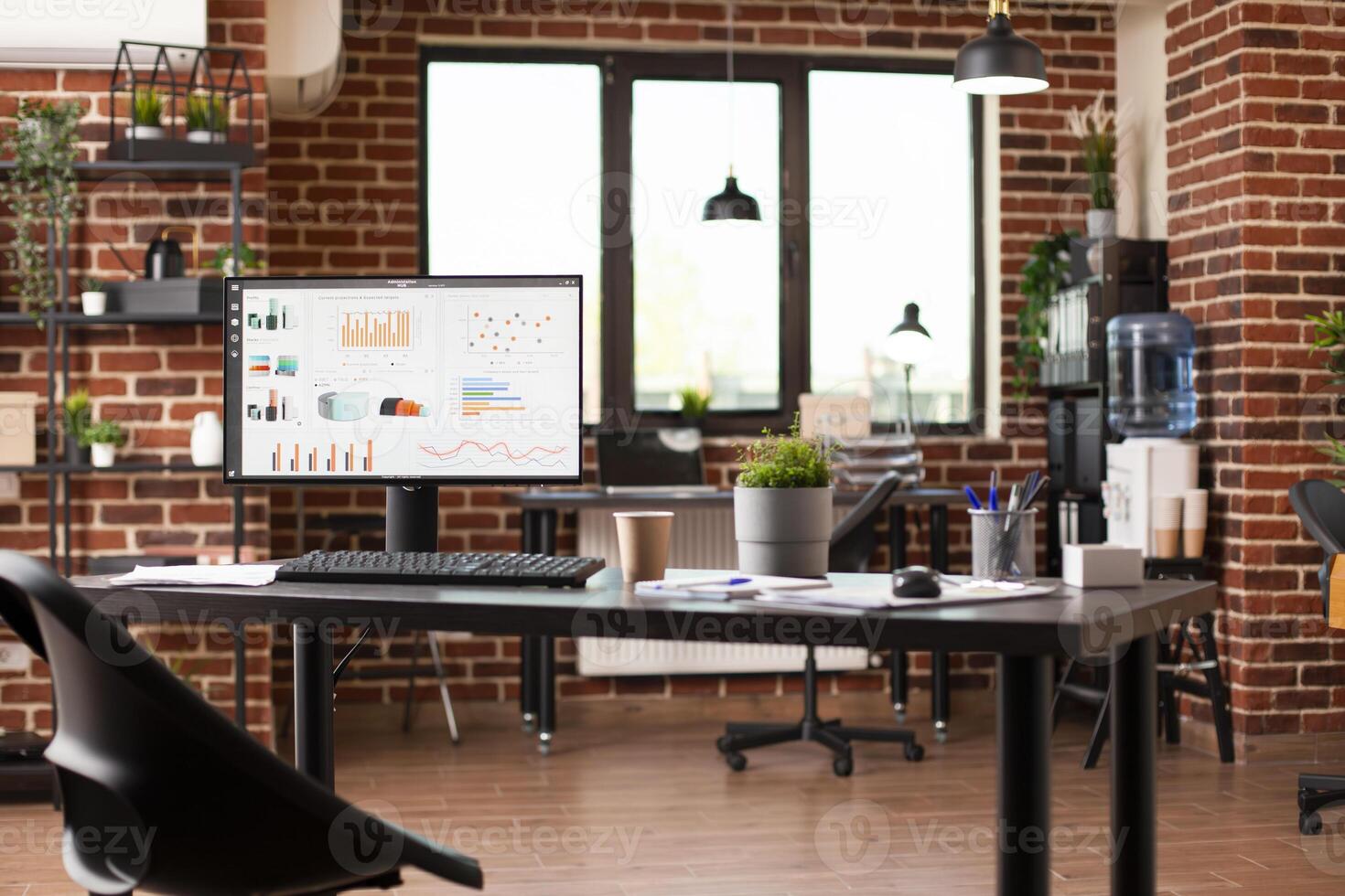 Table in spacious startup office holds desktop pc showing financial analytics. Brick wall interior includes digital devices, shelves, water cooler, and plant decorations, creating refreshing workspace photo