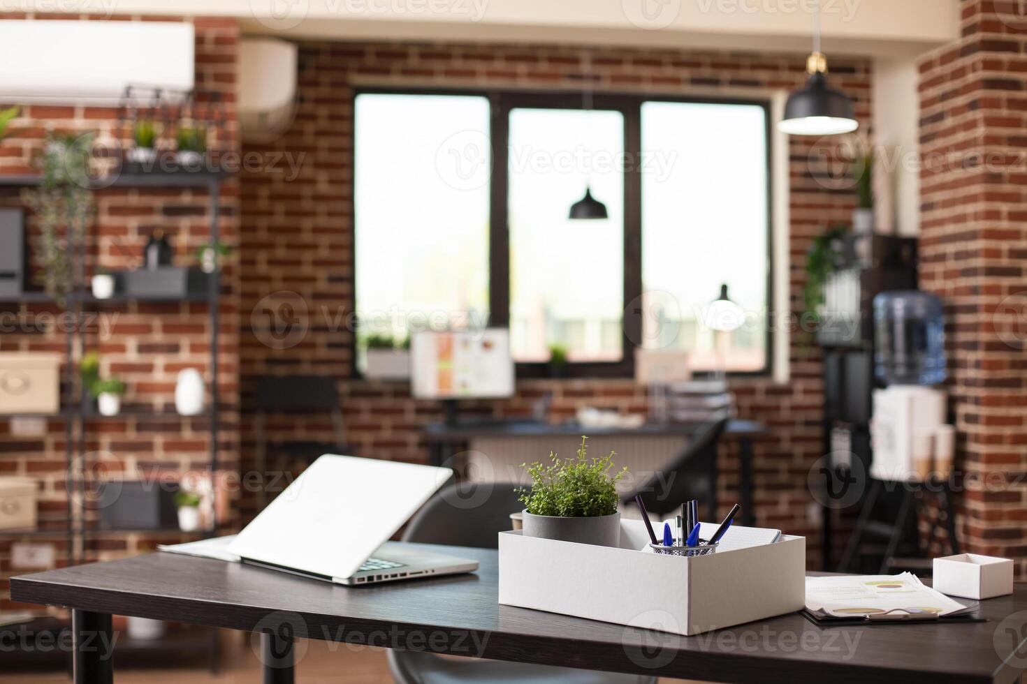 Organized, empty brick wall room with desk, chair, and digital devices for efficient research work at startup company. Clean workspace having a laptop, pot of plant and office materials on table. photo