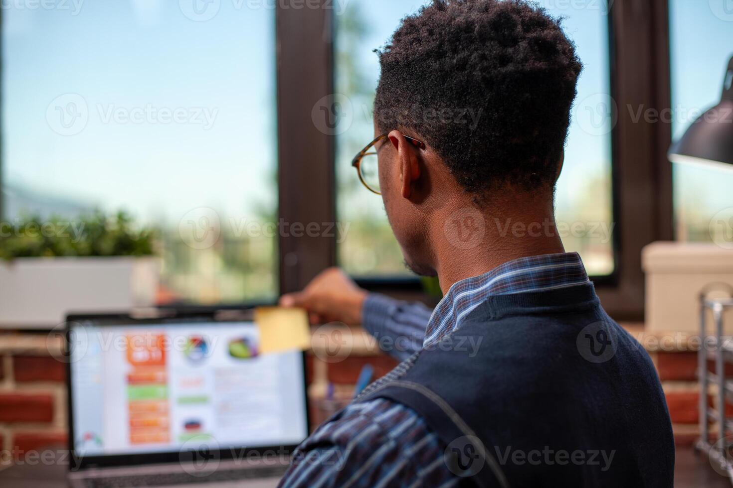 Rearview closeup of african american manager placing a post it on his laptop, preparing for company presentation. Black male researcher sitting at office desk and using sticky notes for reminders. photo