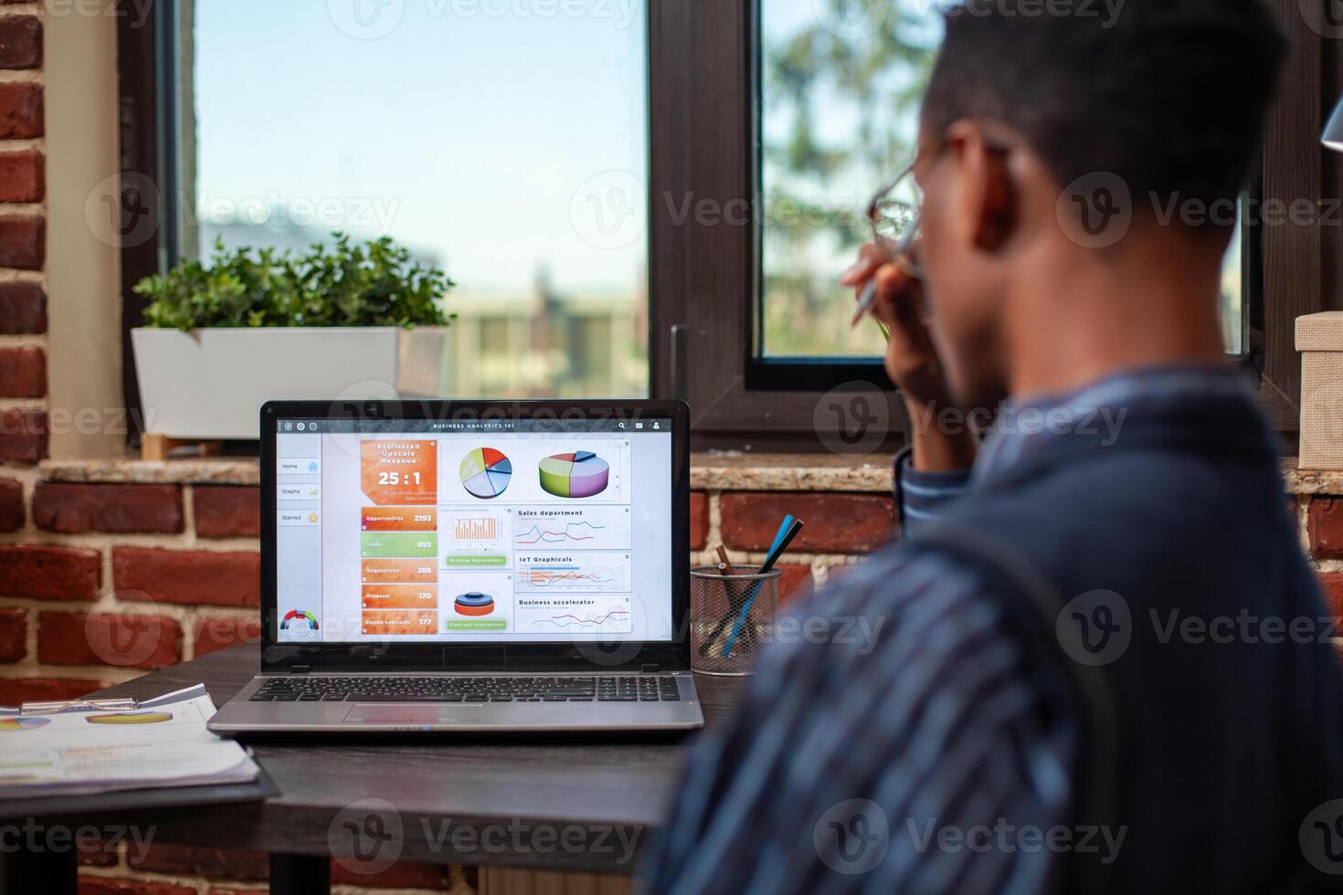 Rearview of african american analyst deeply focused on reviewing business summary analytics displayed on device screen. Male employee looking and reviewing startup project presentation on his laptop. photo