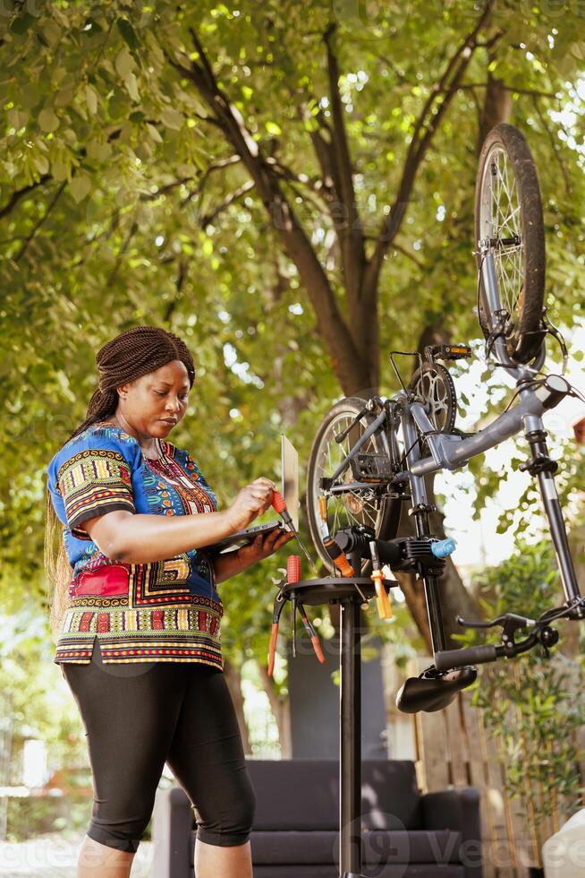 Young healthy active black woman working on modern bicycle by using minicomputer and expert tools. African american female cyclist carrying laptop while searching for specialized equipment in home yard. photo