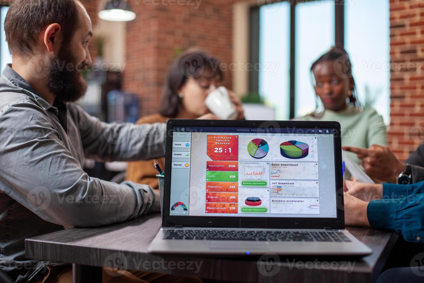 Closeup on device screen showing research analytics as coworkers have a discussion around the table. Laptop displaying marketing strategies is placed on desk at an ongoing meeting of employees. photo
