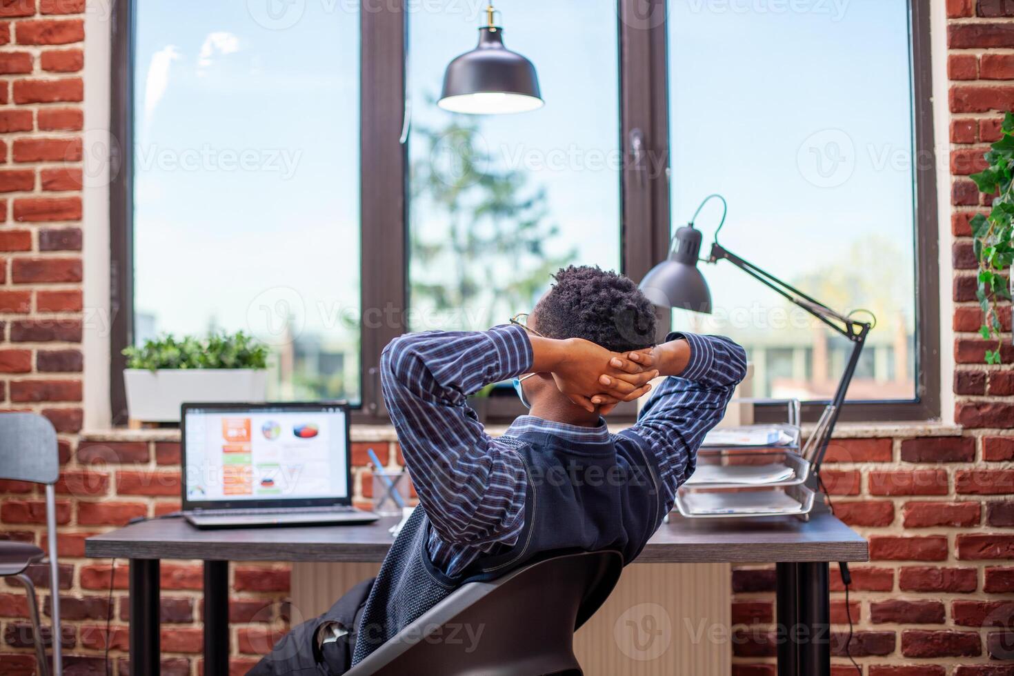 Black entrepreneur stretches his back in modern startup office, feeling accomplished after completing tasks. Male analyst with face mask, leaning in his chair and reflecting on productive day at work. photo