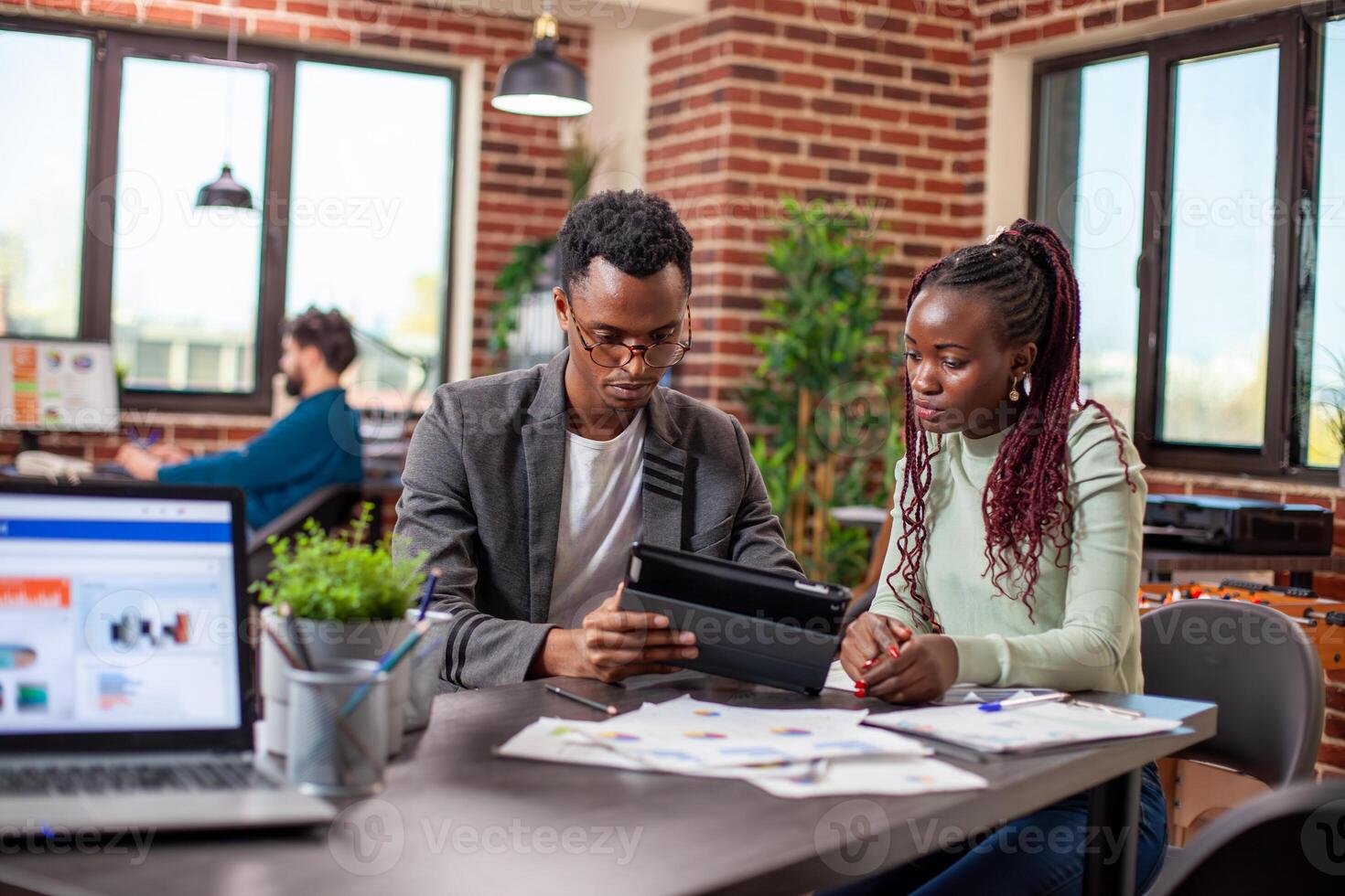 Businessman explaining marketing strategy on tablet to female coworker in brick wall workspace. African american colleagues sitting at table with documents and having a serious conversation in office. photo