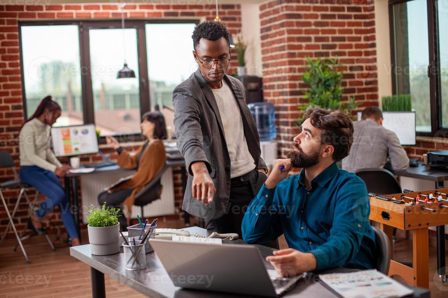 Male analyst points at laptop screen, explaining market analysis update to caucasian coworker. Bearded man sits and listens attentively to black manager describe financial errors in his presentation. photo