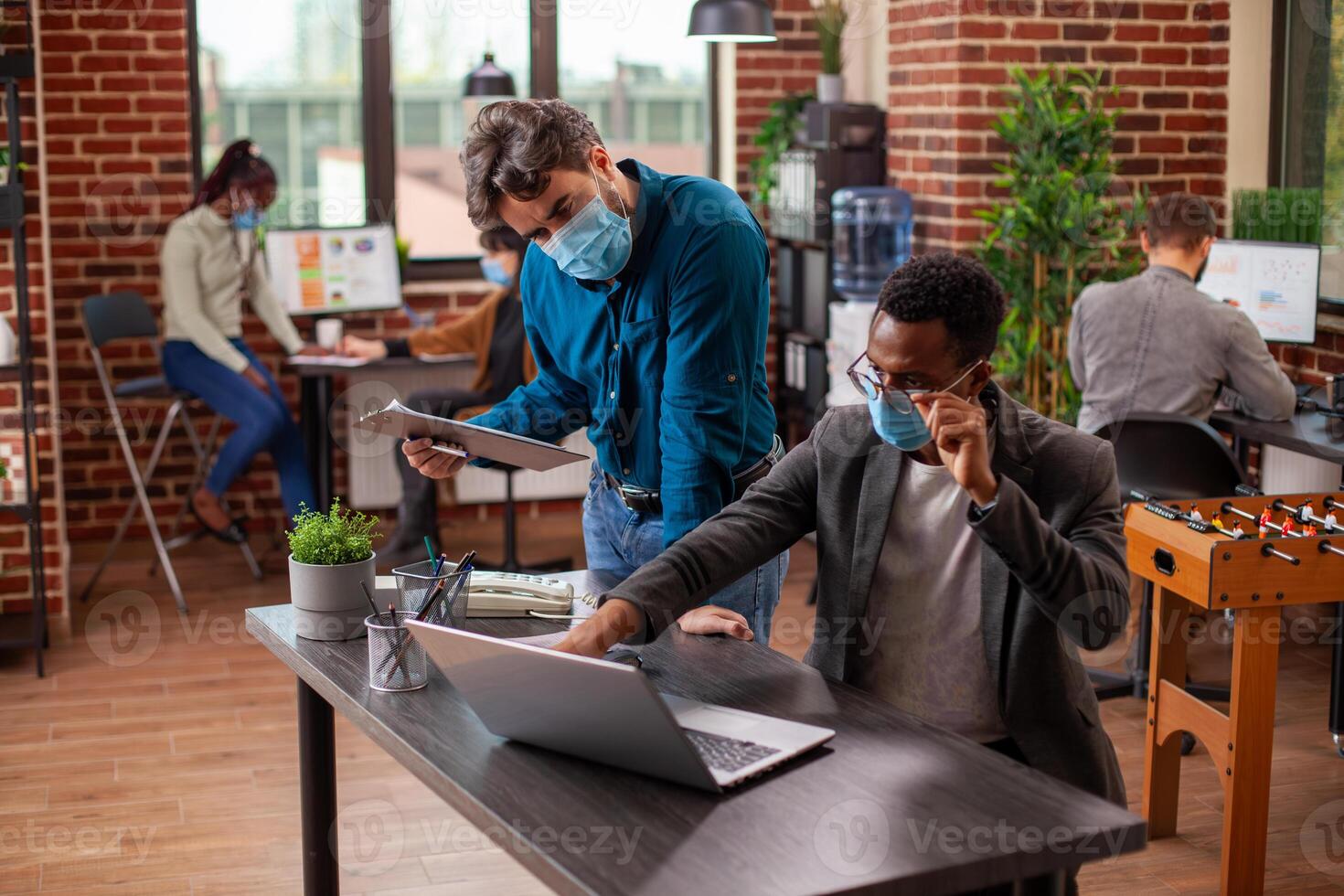 Male professionals discuss business objectives, reviewing documents together on a laptop in modern workspace. Black businessman adjusts his glasses, pointing at device screen and speaking to coworker. photo