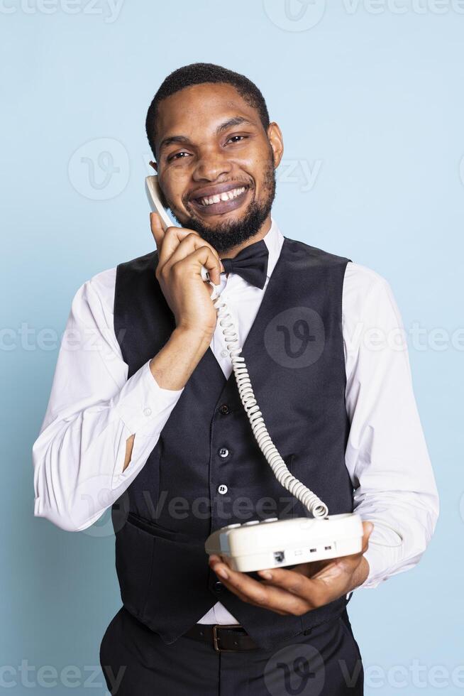 Professional doorkeeper staff makes a room reservation on landline phone call, discussing about hotel amenities and facilities to help guests. Bellboy with suit and tie answers remote call. photo