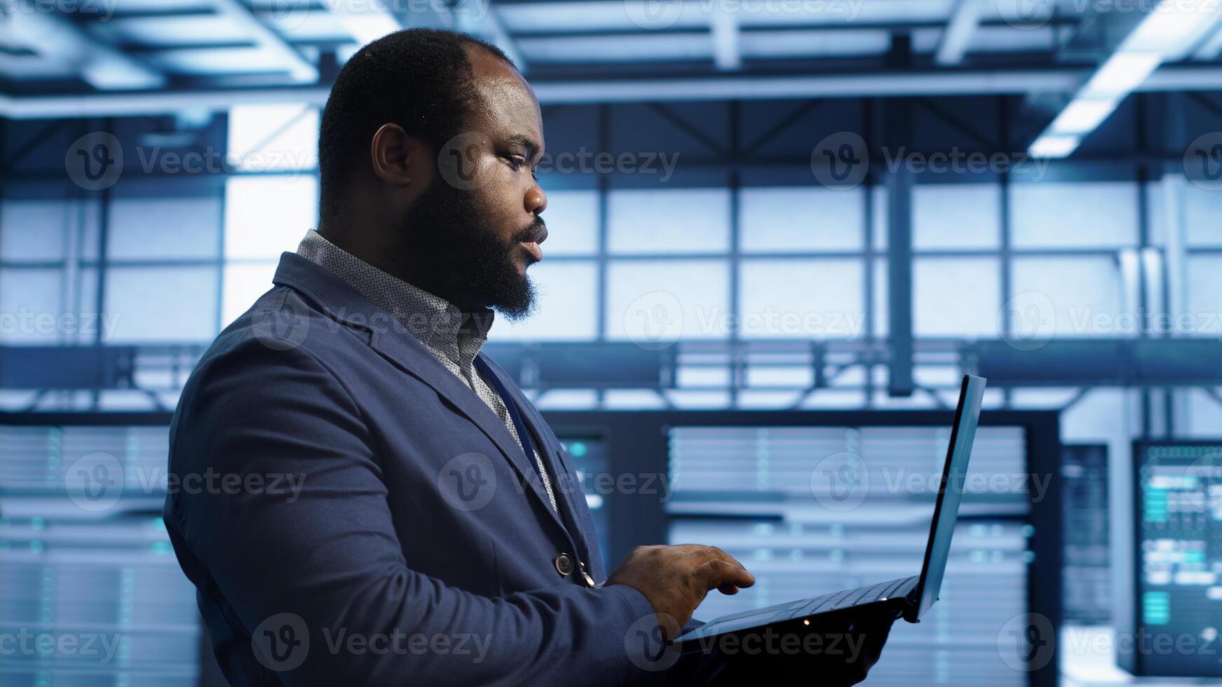 African american man walking in high tech facility, using laptop to patch network issues leading to slow data transfer rates. Data center worker fixing errors affecting supercomputers, camera A photo