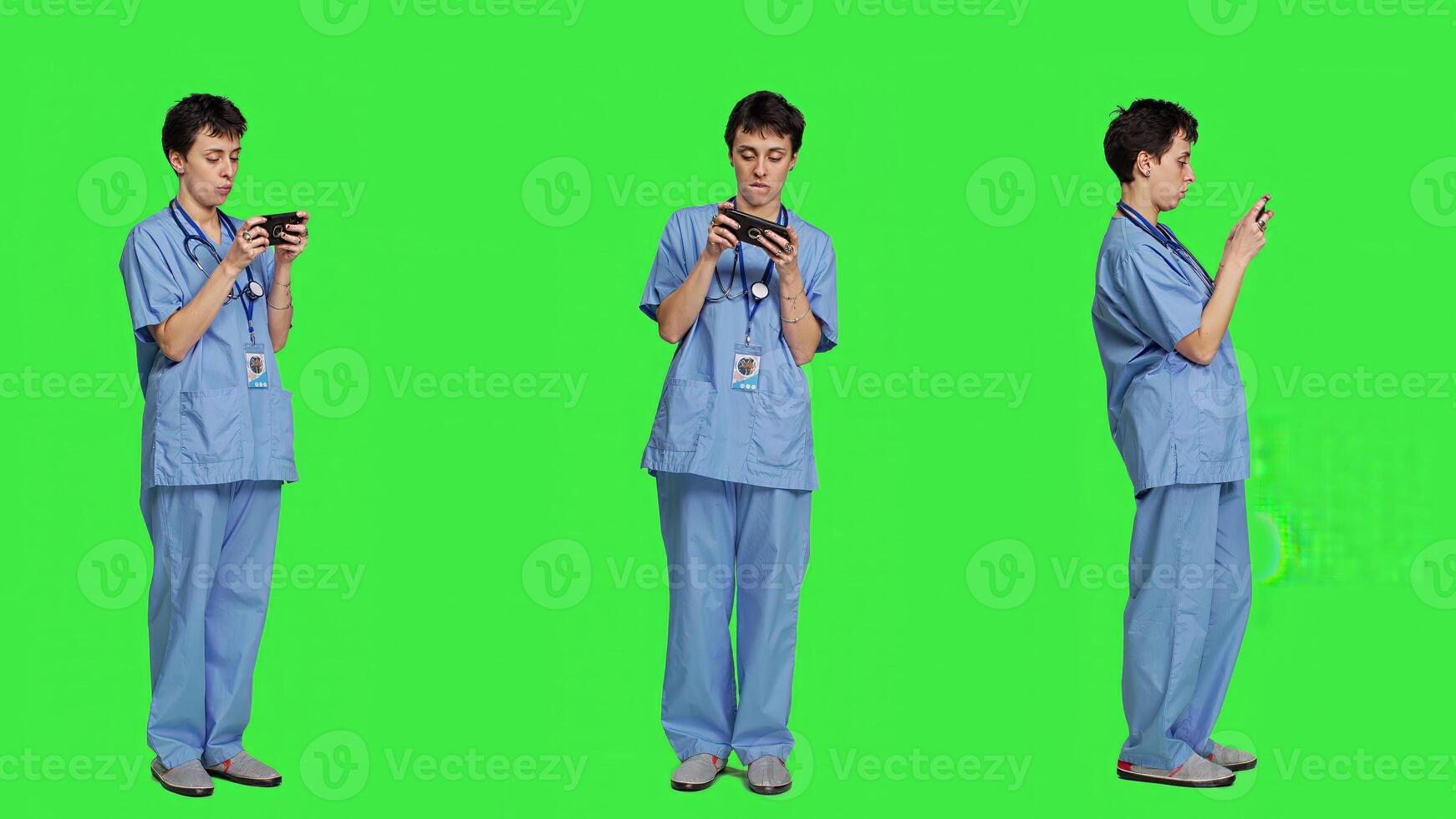 Medical assistant playing mobile games on smartphone app, taking a break from healthcare work. Nurse enjoys gaming tournament on phone, standing against greenscreen backdrop. Camera A. photo