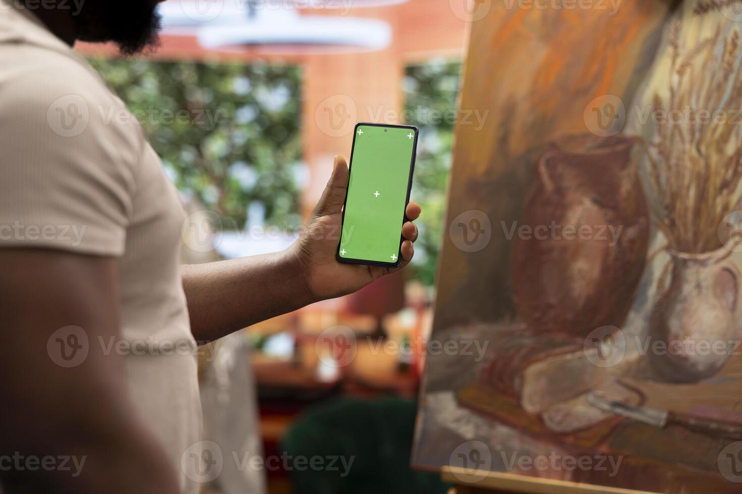 African american artist holding device with green screen mockup in art studio, working on a new painting with a certain technique. Painter using watercolor and acrylic dye for his creation. photo