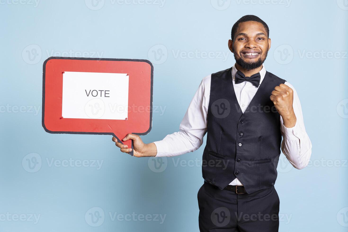 Hotel concierge holding a speech bubble with the message to vote, urging people to get interested in politics on camera. Skilled bellhop with suit and tie showing a marketing ad icon. photo
