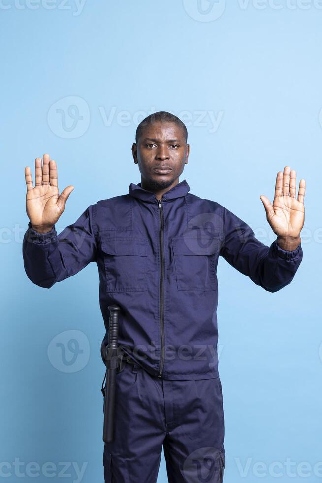 Security agent raising his hands up as a stop symbol in studio, restricting the access with a forbidden sign against blue background. Serious bodyguard in uniform does a limitation gesture. photo