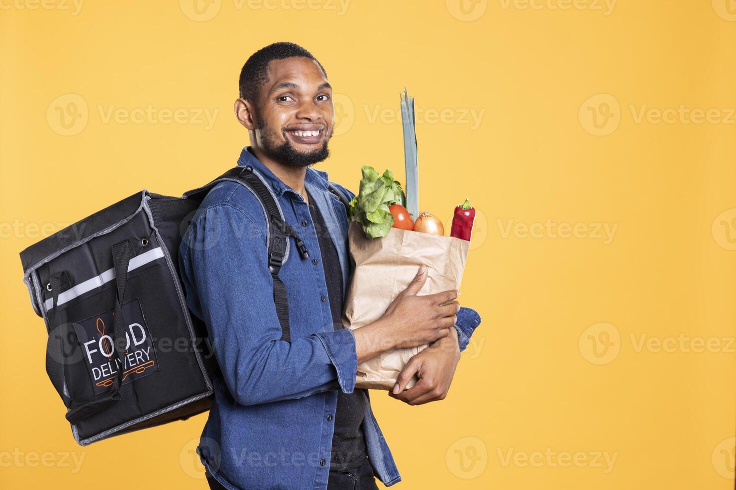 Food delivery courier with a backpack carrying paper bag full of groceries, bringing takeaway food order to customers. African american deliveryman holding fruits and vegetables in studio. photo