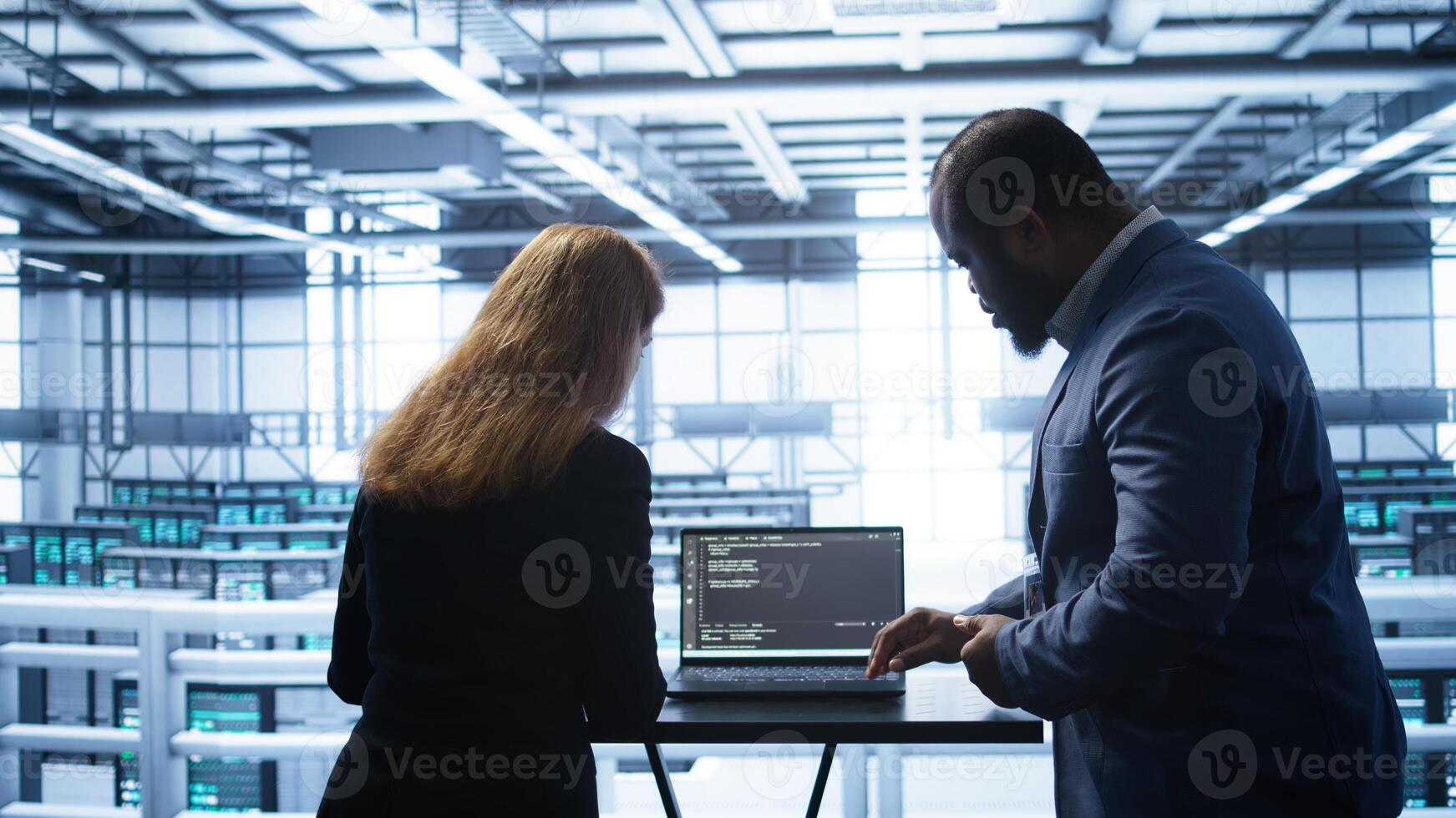 Team of developers doing maintenance checkup using laptop to prolong data center hardware life span. IT support staff members in server hub monitor server infrastructure using notebook, camera B photo