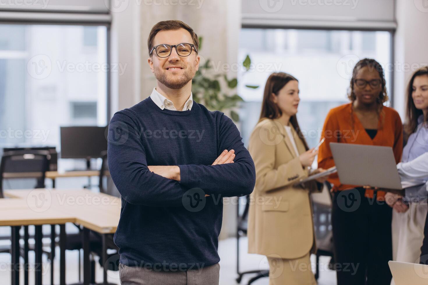 Professional businessman confidently standing with crossed arms, colleagues collaborating in sleek corporate workspace photo