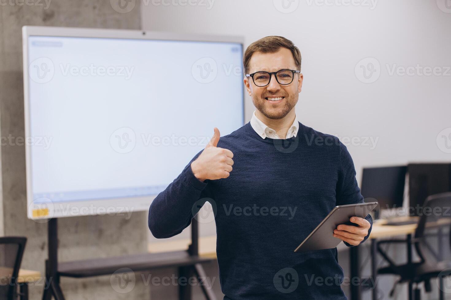 Smiling businessman showing thumbs up while holding tablet, standing in a modern office in front of blank interactive whiteboard photo