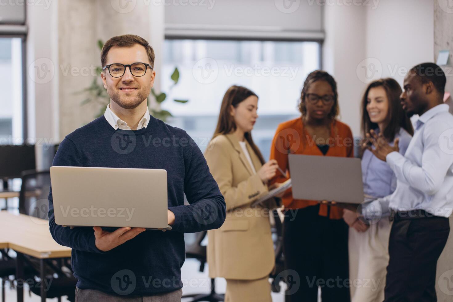 Portrait of smiling businessman holding laptop with diverse business team collaborating on project in modern office photo