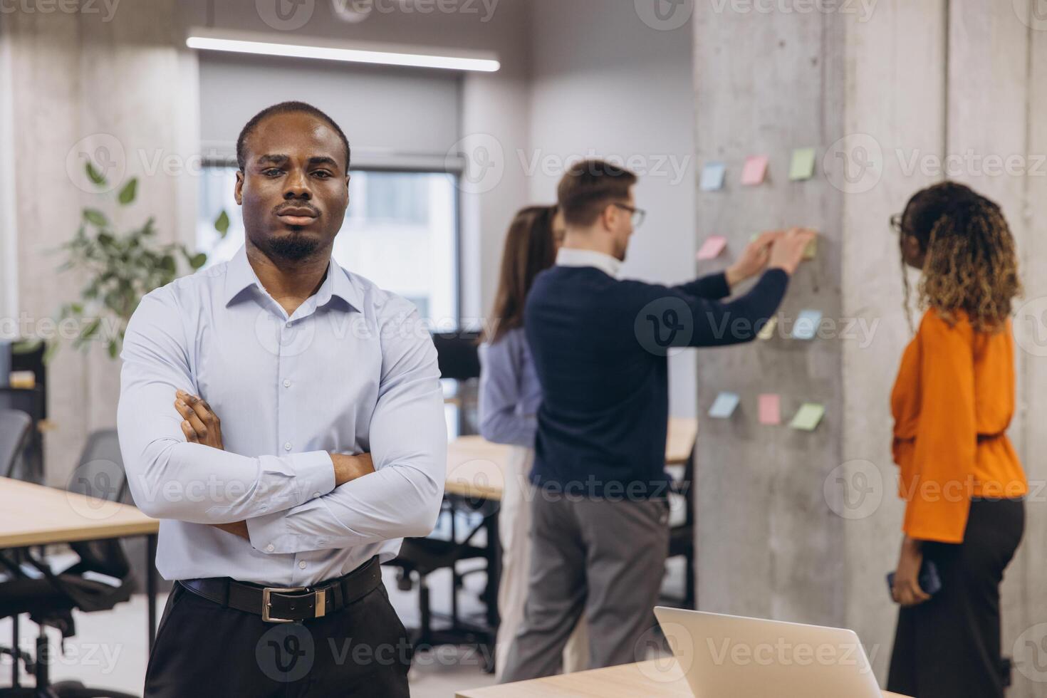 African american male with cross arms, confident professional stands in a contemporary office while colleagues discuss ideas on a board, highlighting teamwork and diversity in business photo