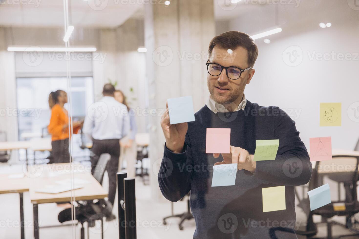 Businessman using sticky notes on a glass wall in a modern office, collaborating with colleagues in the background to brainstorm ideas photo