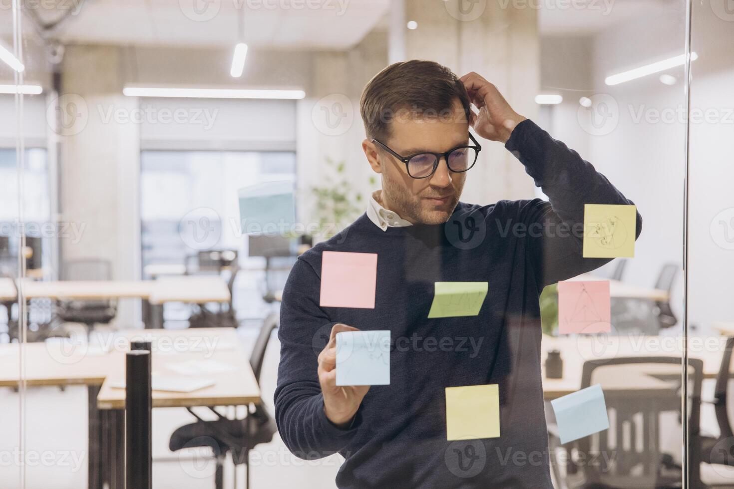 Professional man in office setting brainstorming ideas using colorful sticky notes on a transparent glass wall, focusing on creative solutions photo