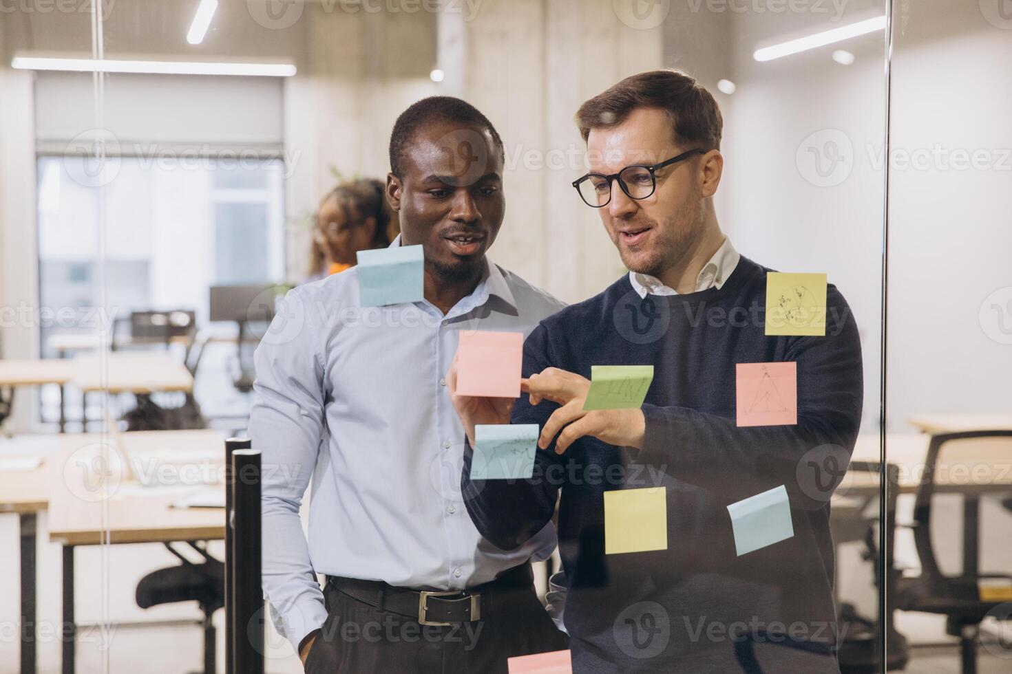 Two businessmen discussing ideas with sticky notes on a glass wall in a modern office, showcasing teamwork and collaboration photo