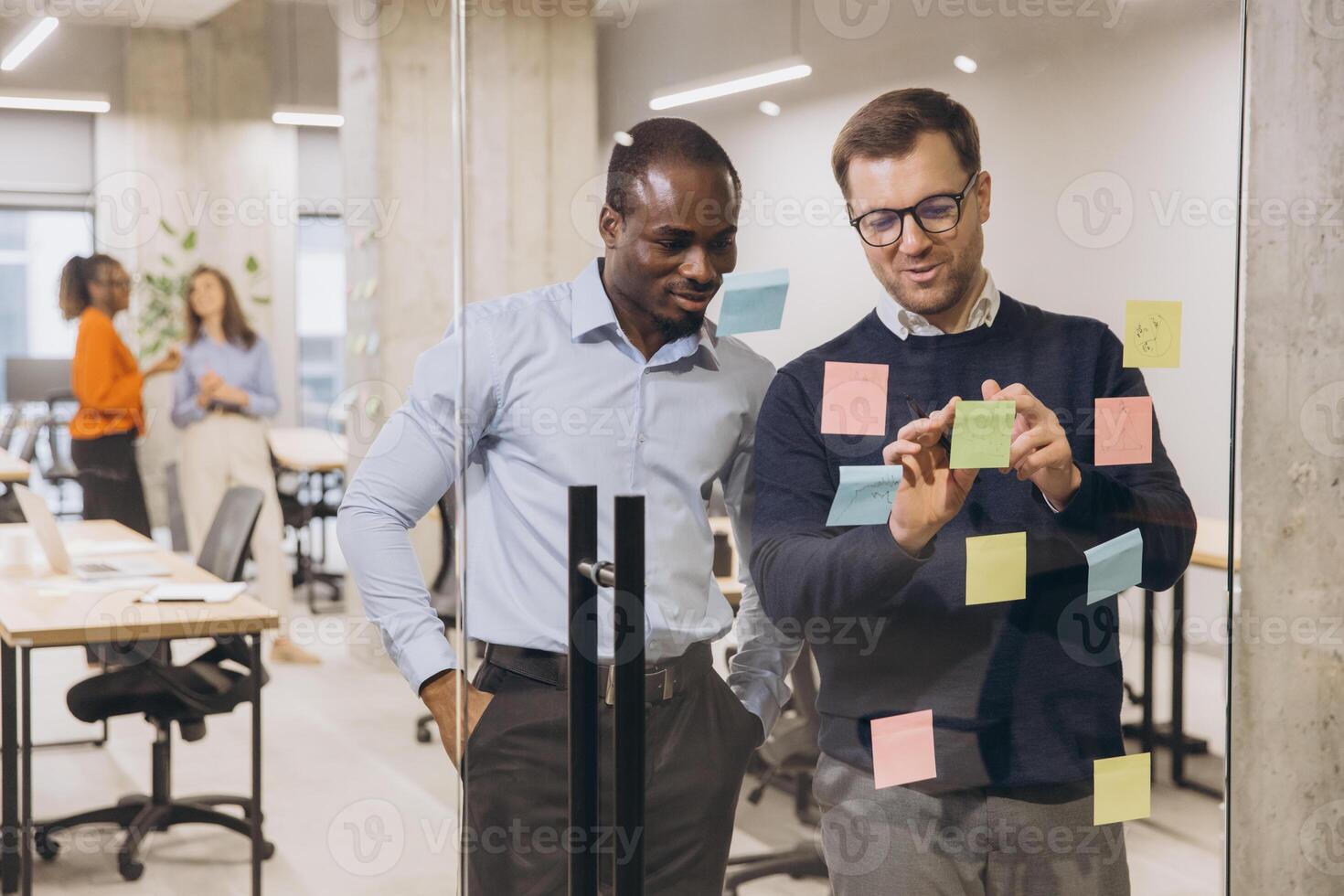 Two diverse businessmen collaborating on a project, using sticky notes on a glass wall in a modern, open plan office setting photo