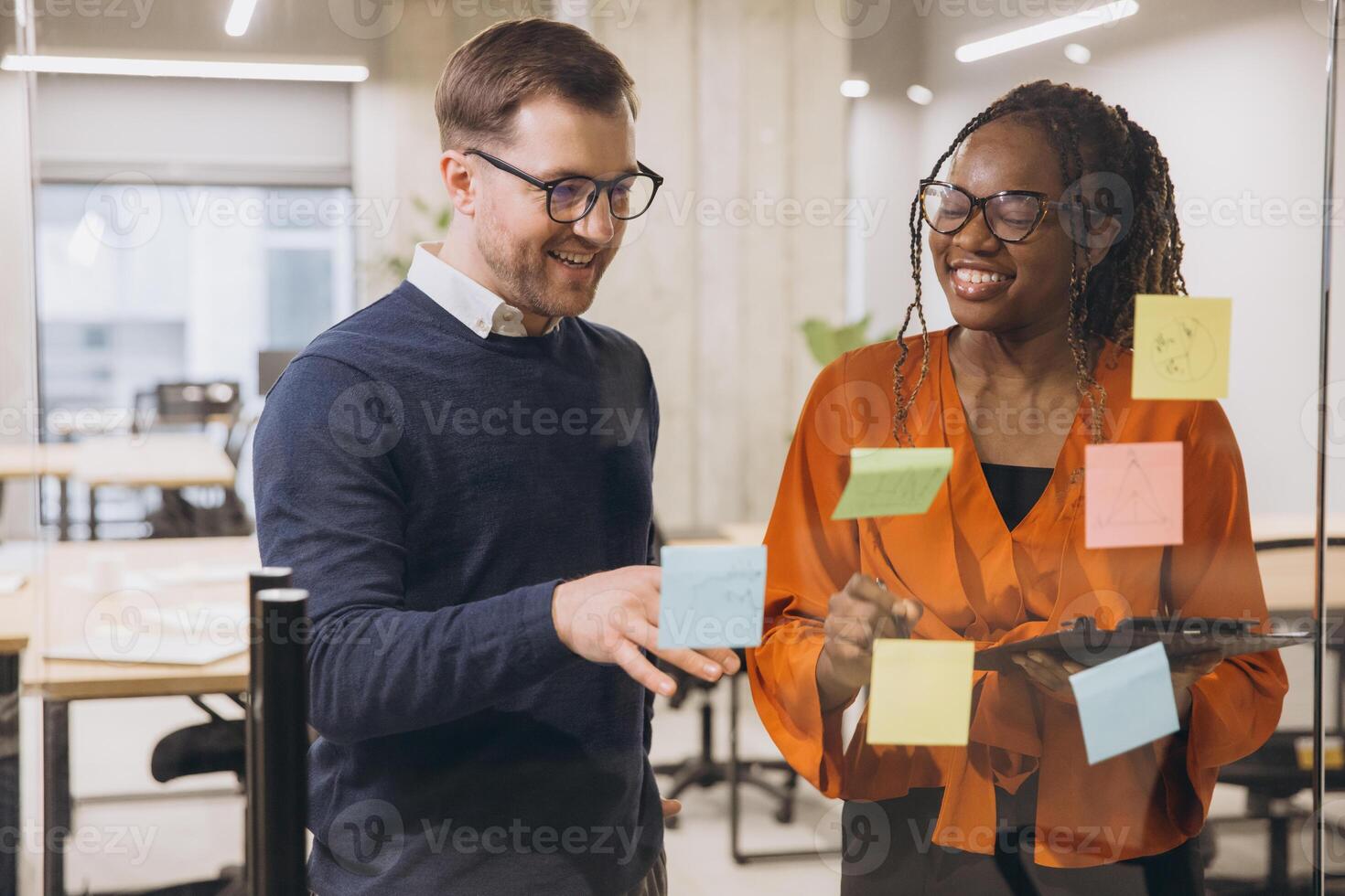 Professional colleagues collaborating, writing ideas on glass wall with sticky notes, demonstrating creative teamwork in modern office environment photo
